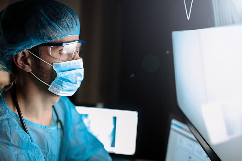 Medical professional in surgical attire, including a mask and protective eyewear, working on a computer screen in a dimly lit room